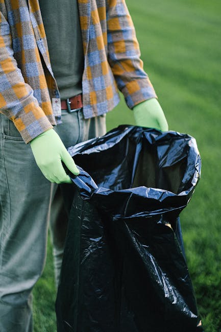 A person wearing a plaid shirt with yellow, gray, and blue tones, paired with gray trousers secured by a brown belt, is standing outdoors on a grassy area. They are holding open a large, black rubbish