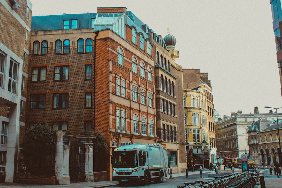 The image depicts a city street scene featuring several multi-story buildings with diverse architectural styles, including ornate brickwork, large windows, and decorative details. A rubbish collection